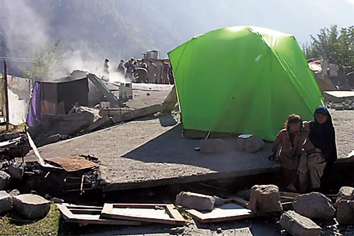 People living in tents set up for evacuation. Collapsed houses remain in the surrounding area.