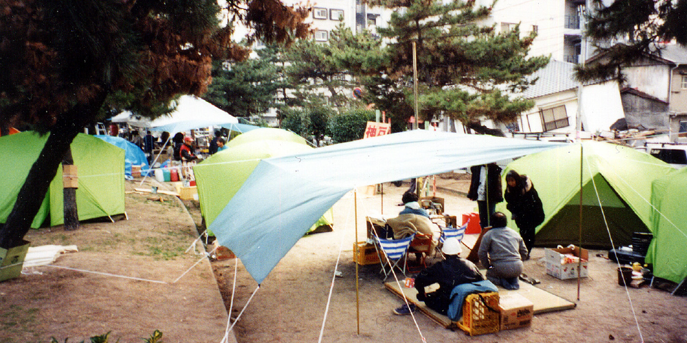 A scene of people living in tents set up in a park after the Great Hanshin-Awaji Earthquake. Collapsed houses remain in the area surrounding the park.