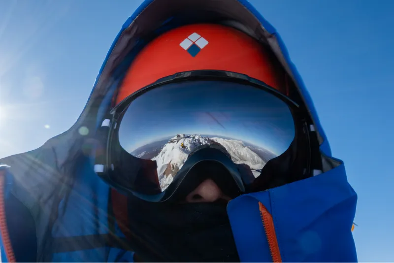 A man wearing blue gear and an orange helmet, with snow-capped mountains reflected in his goggles.