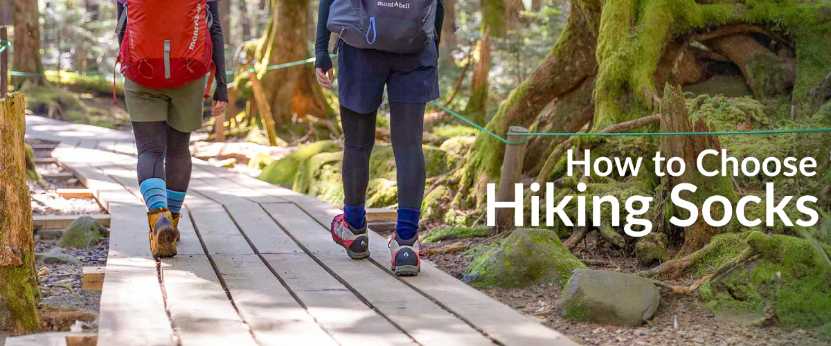 A photo of two women walking along a wooden boardwalk in the forest. The image focuses on their feet, showing them enjoying a trek while wearing light blue hiking socks and gray low-cut socks.