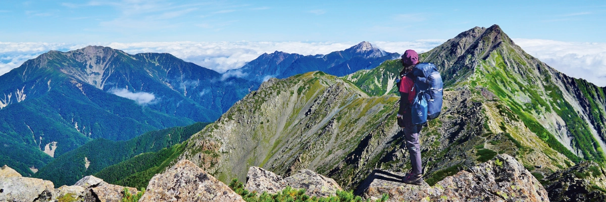A lone climber stands on the ridgeline. Mountain ranges extend into the distance.