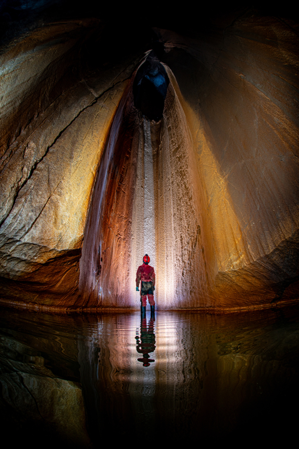 A man wearing a red helmet stands in a cave. His headlamp illuminates the striped patterns on the wall, and the light is also reflected on the water surface at his feet.