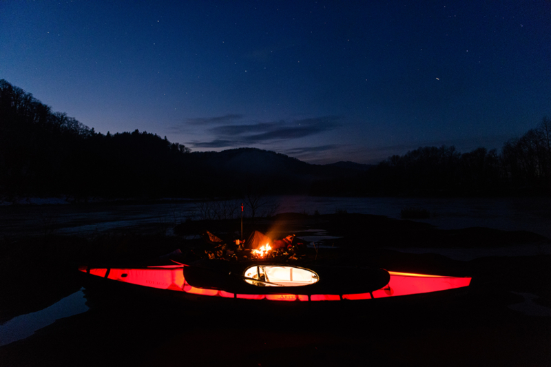 A man lies on his back by the riverside, gazing up at the starry sky. Nearby, a campfire burns, illuminating a red kayak in the foreground.