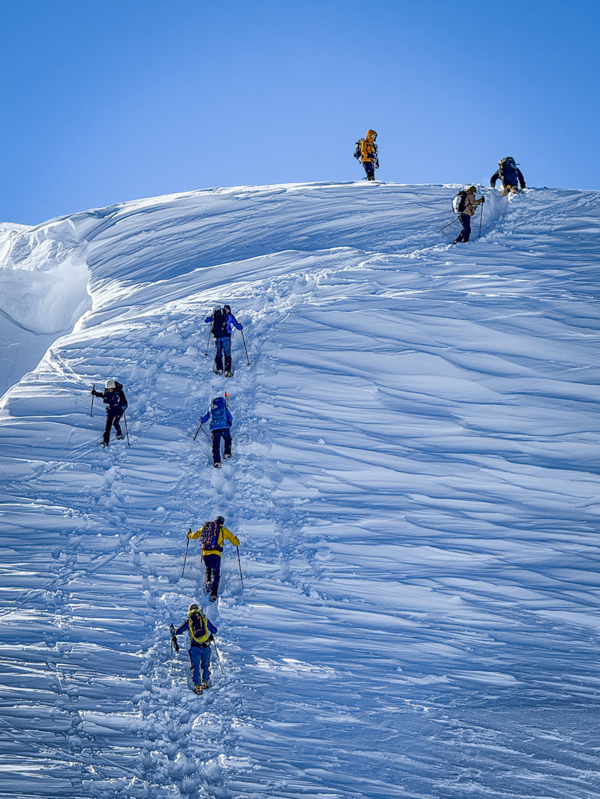 People ascending a steep snow-covered slope. They follow the path trodden by those ahead, climbing in single file.