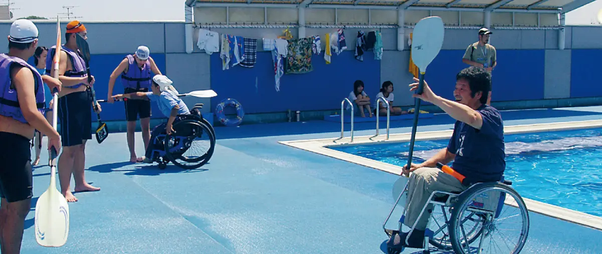 A man in a wheelchair demonstrating paddling techniques with a paddle by the poolside. Several participants are practicing with paddles.