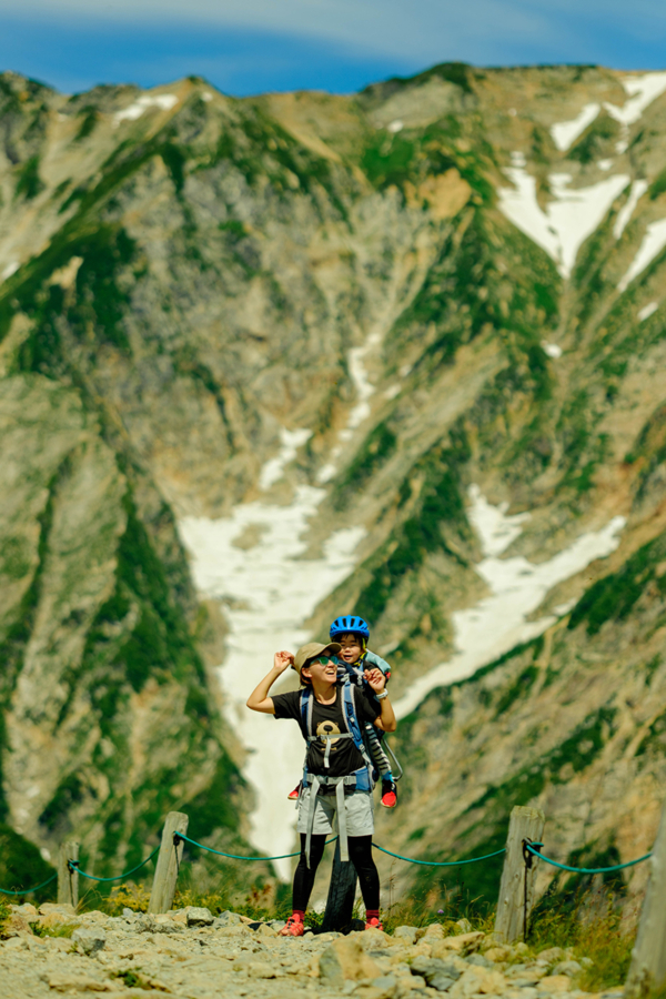 A boy is being carried on his mother's back in a hiking baby carrier. Behind the smiling pair, a majestic mountain with remnants of snow towers in the background.