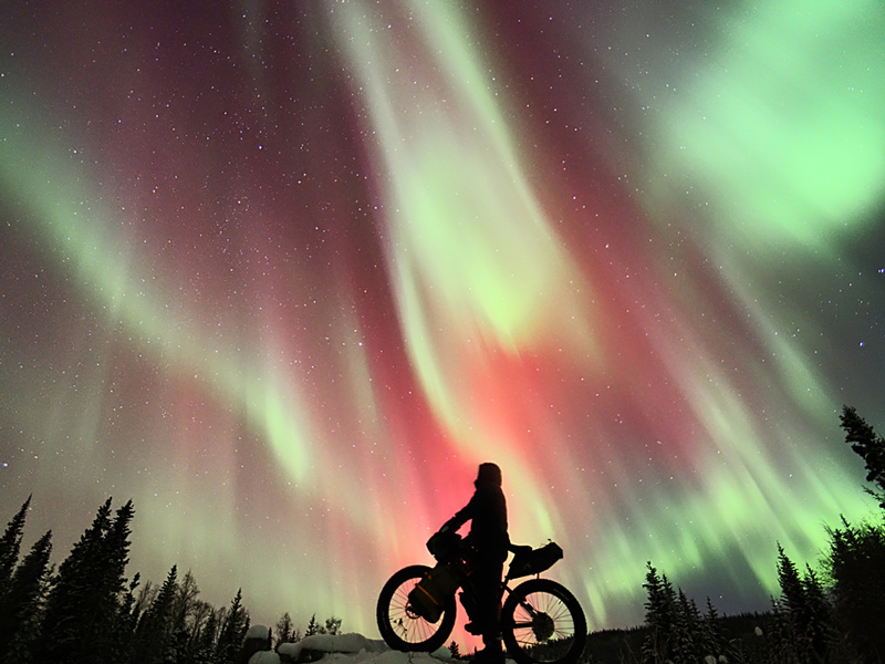 A cyclist gazing up at red and green aurora borealis over their bicycle