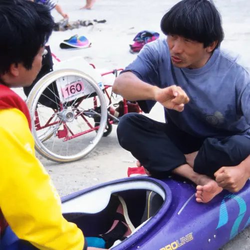 Isamu Tatsuno demonstrates kayaking techniques and teaches paddling to a wheelchair user with a disability.