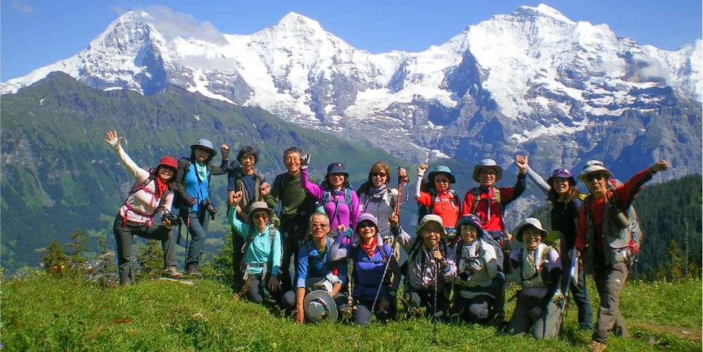 A group photo of about a dozen people enjoying trekking. In the background, snow-capped mountain ridges stretch across the horizon.