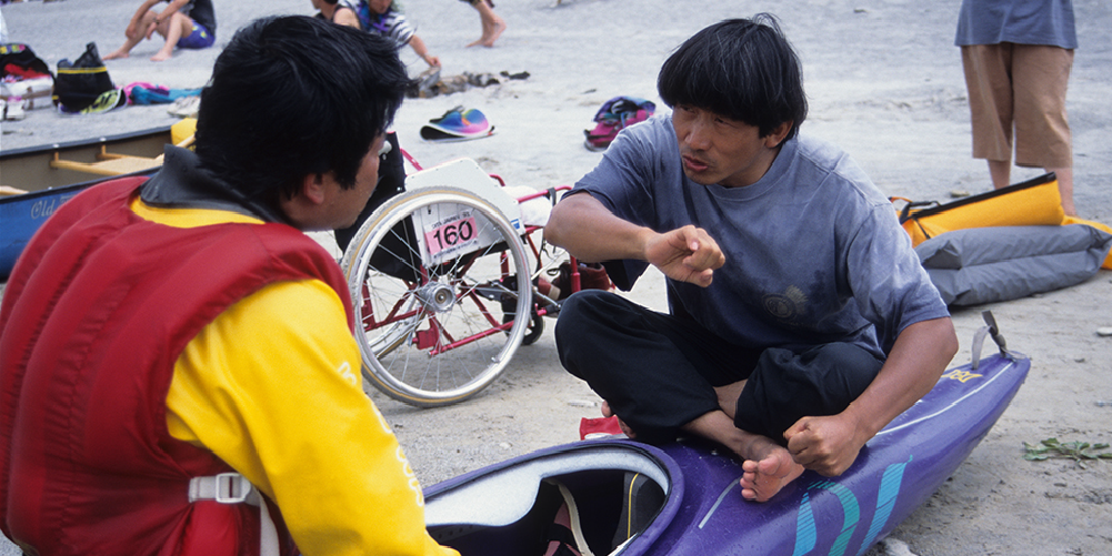 Isamu Tatsuno demonstrates proper kayak paddling technique and teaches a person with disabilities in a wheelchair how to paddle.