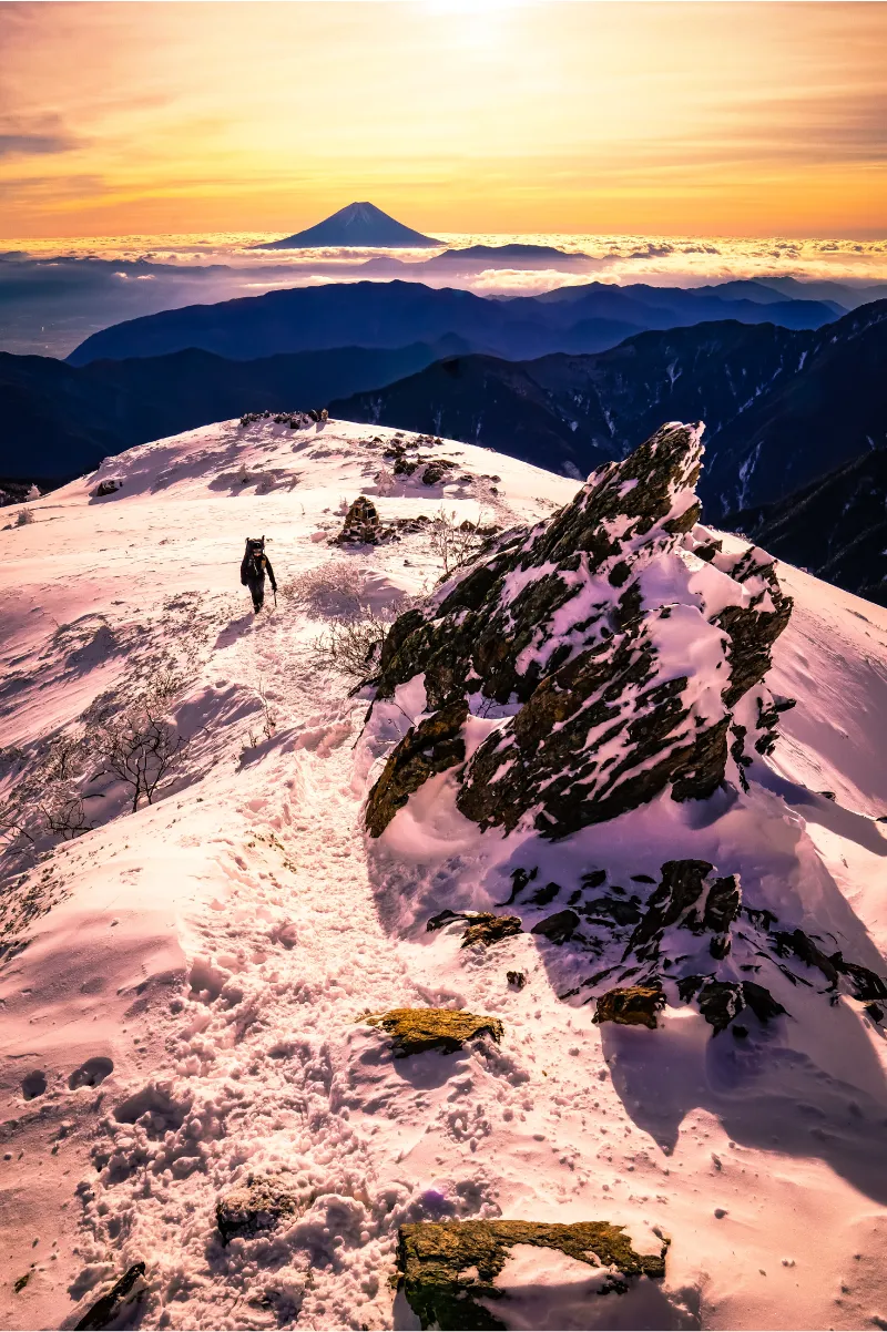 A man with an ice axe is walking along a snow-covered mountain ridge, with Mount Fuji visible in the distance floating above a sea of clouds.
