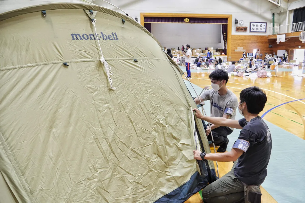 Two Outdoor Gientai staff members setting up tents in an evacuation center gymnasium.