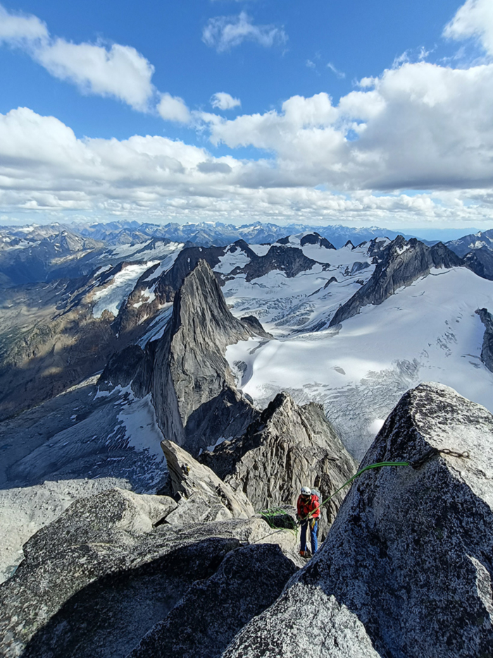 Mountains extend endlessly towards the horizon. From a large rock face in the foreground, a single rope hangs down, and a climber in a red jacket is rappelling down.