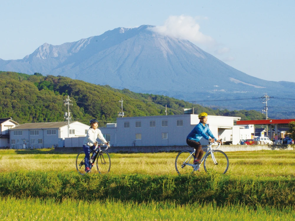 Two women are cycling along a path between rice paddies